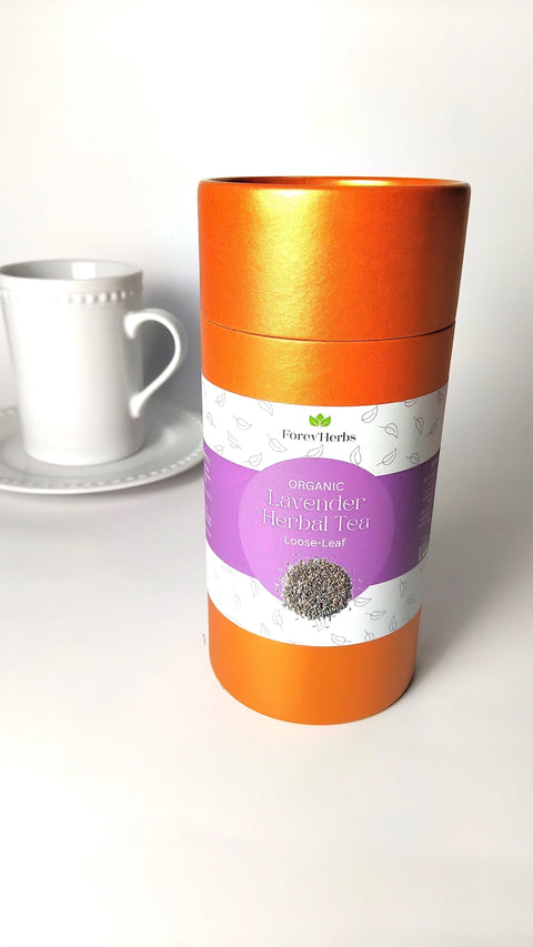 A tall orange canister of Forev'Herbs Organic Lavender Herbal Tea stands in front of a white teacup and saucer on a white surface with a plain white background, ideal for enjoying this calming herbal blend.