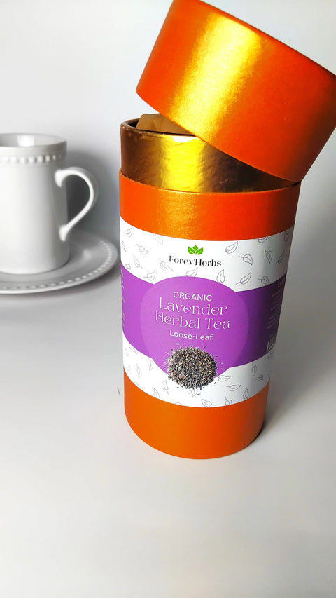 An open cylindrical orange and gold Forev'Herbs container labeled "Organic Lavender Herbal Tea Loose Leaf" sits with its tilted lid; a white cup and saucer are visible in the background.