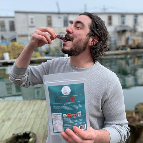 A dark-haired, bearded man smiles while eating Oceans Balance Organic Whole Leaf Dulse, holding the package on a wooden dock with water, lobster traps, and buildings in the background.