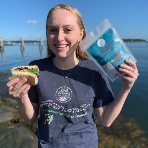 A smiling woman stands by the water, holding a sandwich and an Organic Whole Leaf Dulse package from Oceans Balance. She wears a navy Oceans Balance t-shirt, with wooden posts and blue sky in the background.