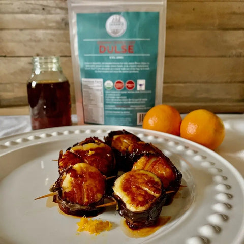 A white plate with bacon-wrapped scallops garnished with orange zest sits beside two mandarins, a jar of dark liquid, and a package of Oceans Balance Organic Whole Leaf Dulse, spotlighting this superfood in the background.