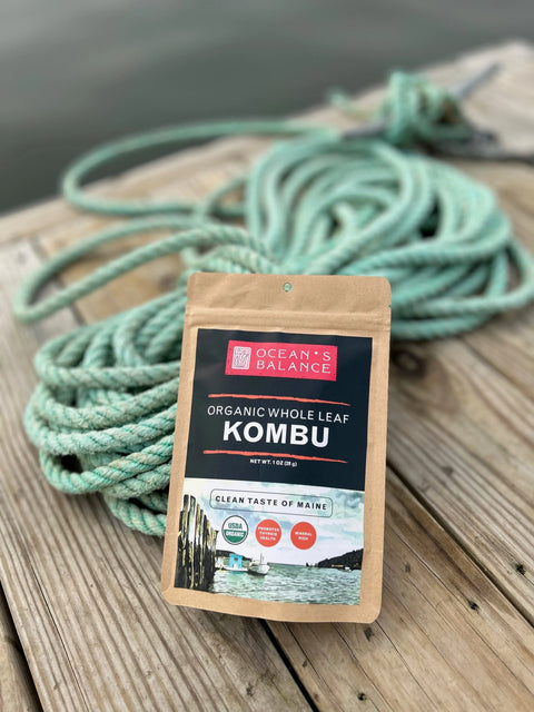 A package of Oceans Balance Organic Whole Leaf Kombu sits on wooden planks by a coiled green rope, with water in the background—ideal for making dashi or savoring authentic organic seaweed flavor.