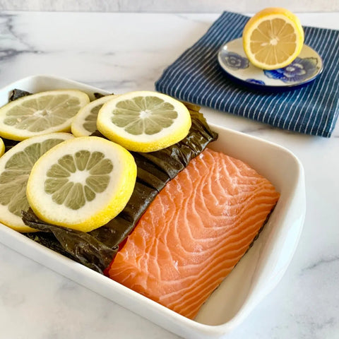 A raw salmon fillet in a white dish is partially covered with Oceans Balance Organic Whole Leaf Kombu and topped with lemon slices. In the background, a blue-striped napkin holds a plate with a halved lemon.