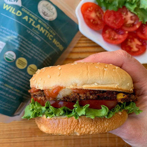 A hand holds a sandwich with lettuce, tomato, and a crispy patty in a sesame bun. In the background are Oceans Balance Organic Wild Atlantic Nori Whole Leaf and a plate of sliced tomatoes and lettuce.