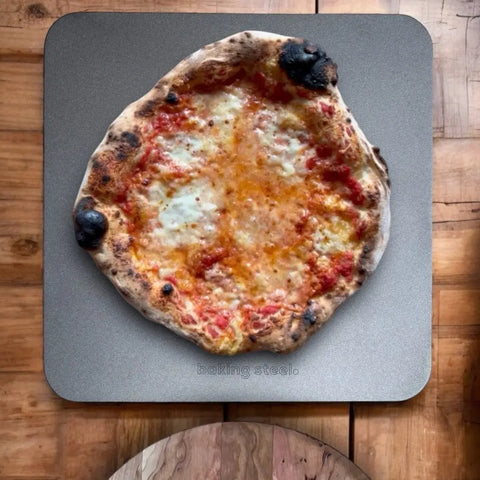 A rustic margherita pizza with melted cheese and tomato sauce rests on a Baking Steel Original by Baking Steel ® atop a wooden table. The charred crust edges highlight the steels superior performance.