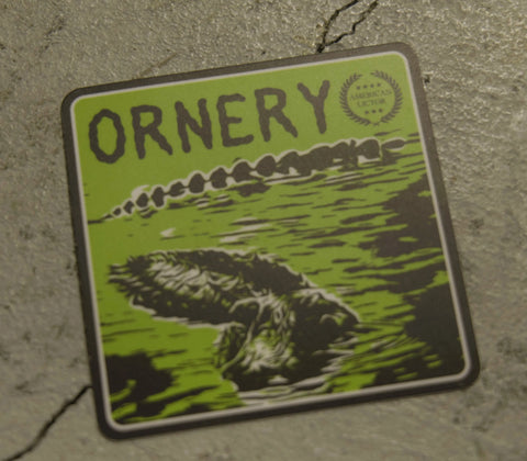 The Sticker: Alligator - Ornery by American Lictor is a green sticker with ORNERY at the top, an alligator in water suggesting armored muscle, and a small American Lictor seal in the corner, shown on a textured surface.