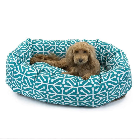 A small brown dog rests on the Majestic Pet Aruba Bagel Dog Bed, featuring a blue-and-white geometric bone pattern and machine washable cover, set against a plain white background.