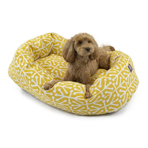 A small brown curly-haired dog relaxes in the Majestic Pet Aruba Bagel Dog Bed, featuring a yellow color with a white bone pattern. The cozy bed is machine washable for easy care.