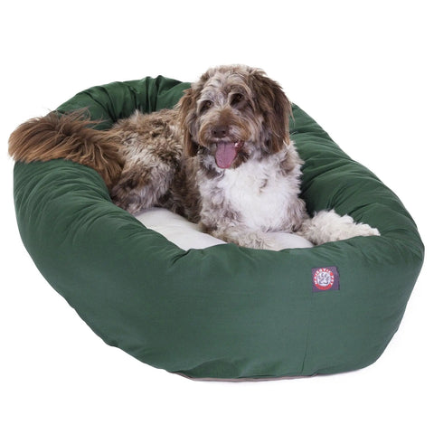 A brown and white, long-haired dog lies on a green Poly/Cotton Sherpa Bagel Dog Bed by Majestic Pet, looking at the camera with its tongue out.