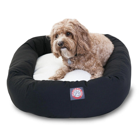 A small, curly-haired brown dog lounges in a Majestic Pet Poly/Cotton Sherpa Bagel Dog Bed, featuring a round black and white design with a cozy sherpa cushion and water-resistant fabric.
