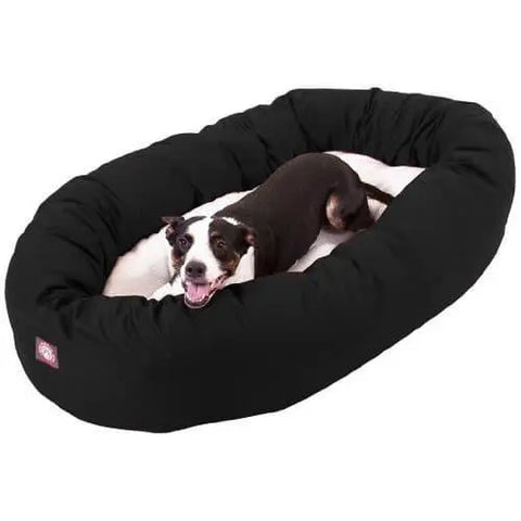 A black and white dog lies happily in a large Majestic Pet Poly/Cotton Sherpa Bagel Dog Bed, looking up with its mouth open and tongue out. The bed is machine washable for easy cleaning.