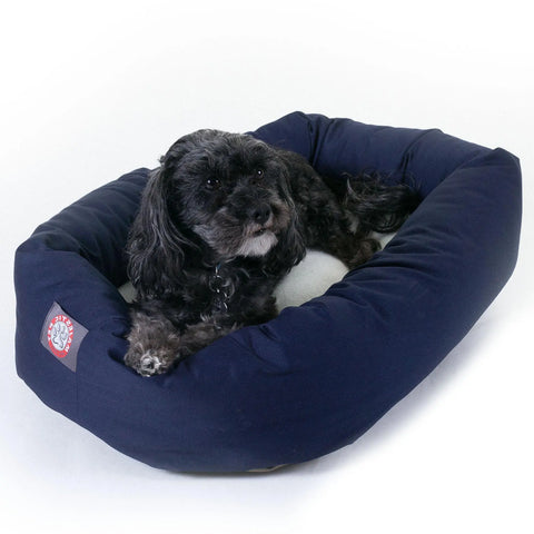 A small black dog with curly fur relaxes in a navy blue Majestic Pet Poly/Cotton Sherpa Bagel Dog Bed with a white sherpa cushion, looking off to the side.