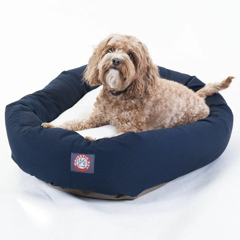 A small, curly-haired brown dog lies on a Majestic Pet Poly/Cotton Sherpa Bagel Dog Bed in navy blue with a white cushion and red-and-white brand patch. The cozy bed is machine washable and set against a plain white background.