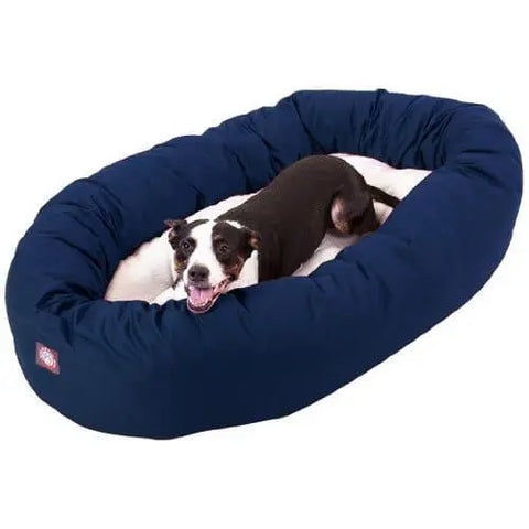 A black and white dog relaxes in the Majestic Pet Poly/Cotton Sherpa Bagel Dog Bed, featuring a navy blue, water-resistant, oval design with a plush Sherpa cushion. The dog looks up happily with its mouth open and tongue out.