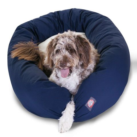 A fluffy brown and white dog sits comfortably in a large, navy Poly/Cotton Sherpa Bagel Dog Bed by Majestic Pet, looking at the camera with its tongue out. One front leg rests on the edge of the water-resistant bed.
