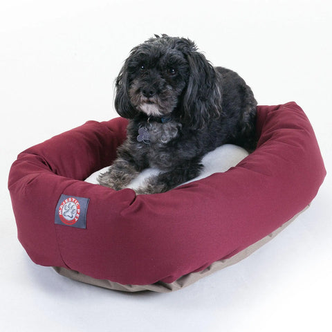 A small black and gray dog with curly fur lies on a maroon and tan Majestic Pet Poly/Cotton Sherpa Bagel Dog Bed, looking toward the camera against a plain white background.