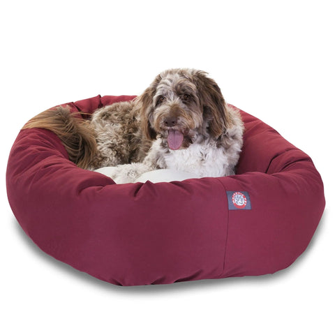 A fluffy brown and white dog relaxes in the Majestic Pet Poly/Cotton Sherpa Bagel Dog Bed with a soft cushion, looking at the camera with its tongue out. The bed is maroon, and the background is plain white.