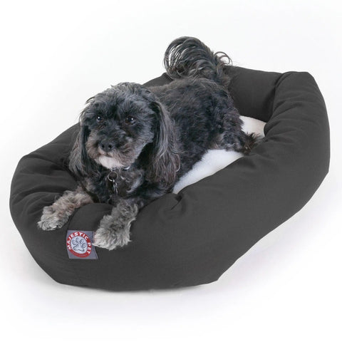 A small black and gray dog with curly fur is lying on a Majestic Pet Poly/Cotton Sherpa Bagel Dog Bed, looking up at the camera.