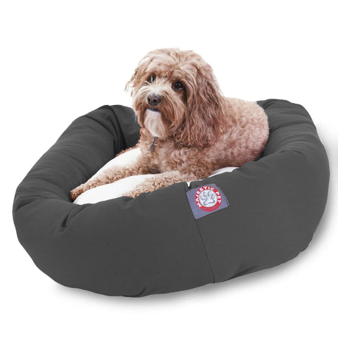 A small, curly-haired dog with light brown fur is lying on a Majestic Pet Poly/Cotton Sherpa Bagel Dog Bed. The cozy bed features a logo patch and has a machine washable cover, making it easy to keep clean while your dog relaxes.