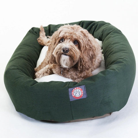 A small, curly-haired brown dog relaxes on the white Sherpa cushion of a plush, dark green Poly/Cotton Sherpa Bagel Dog Bed by Majestic Pet, adding charm to the cozy scene.