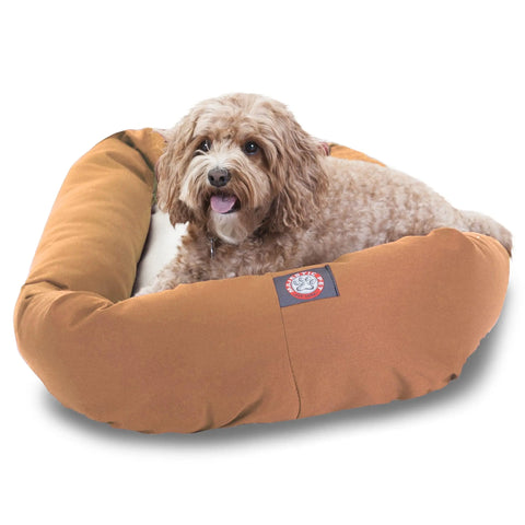 A small, curly-haired brown dog relaxes in a Majestic Pet Poly/Cotton Sherpa Bagel Dog Bed, enjoying its plush, water-resistant comfort and cozy sherpa cushion with its tongue slightly out.