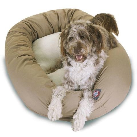 A shaggy brown and white dog lounges in the Majestic Pet Poly/Cotton Sherpa Bagel Dog Bed, a round tan bed with a plush Sherpa cushion, resting its front paws on the edge and gazing upward with an open mouth.