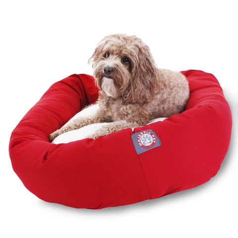 A small, curly-haired brown dog lies on a Majestic Pet Poly/Cotton Sherpa Bagel Dog Bed with a plush red, water-resistant cover and soft sherpa cushion, looking up. The bed displays a paw print logo label on the side.