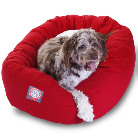 A fluffy brown and white dog lounges on a bright red Majestic Pet Poly/Cotton Sherpa Bagel Dog Bed, looking at the camera with its mouth open and tongue out.