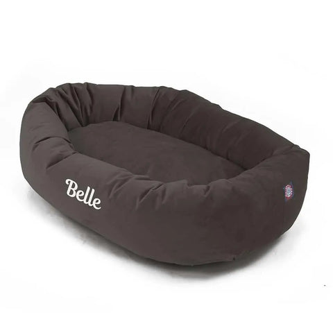 The Majestic Pet Velvet Bagel Dog Bed is a dark brown, oval, high-sided bed with Belle in white on the front and a brand logo on the right. Personalize it for your pets comfort and style.