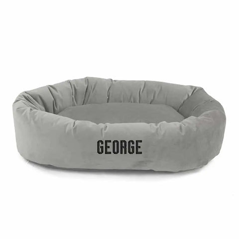 The Majestic Pet Velvet Bagel Dog Bed - Personalized is a light gray, soft round bed with “GEORGE” embroidered in black on the front. It features raised sides, a cushioned base, and gentle spine support for your pet.