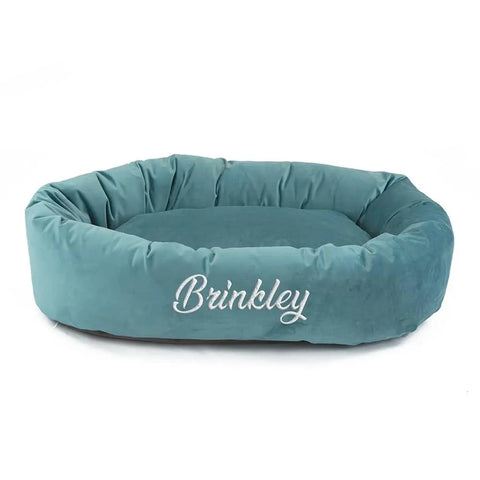 The Majestic Pet Velvet Bagel Dog Bed - Personalized in teal features an oval shape with Brinkley embroidered in white. It has plush, raised sides and a cushioned bottom for comfort, plus its machine washable for easy cleaning.