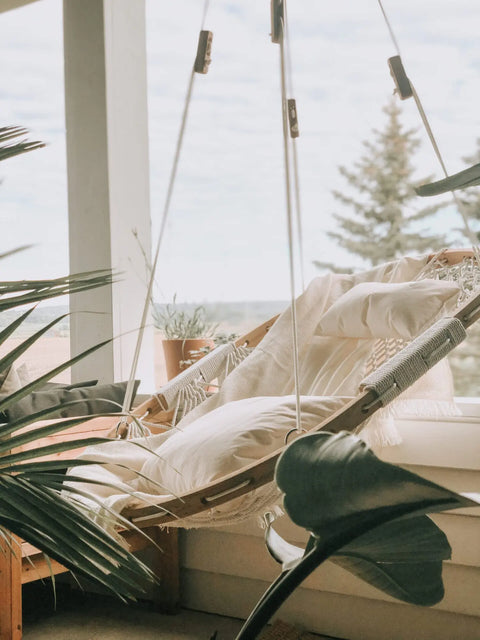 A Cobble Mountain Replacement Pillow/Cushion in white rests on a cozy hammock chair on the porch, surrounded by greenery and large windows framing views of trees and a bright, cloudy sky.