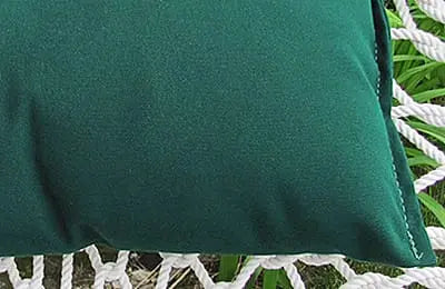 A close-up of a Cobble Mountain Replacement Pillow in dark green, designed for outdoor use, rests on a white woven hammock with lush green plants in the background.