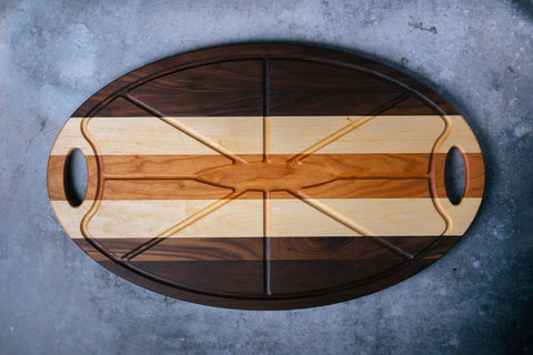 The Oval Carver by Montana Block is a handmade solid wood cutting board with dark and light stripes, engraved grooves, and dual handles, shown on a gray textured surface.