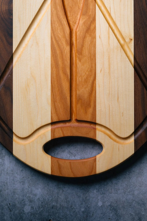The Oval Carver by Montana Block is a handmade solid wood cutting board with alternating light and dark stripes, a carved groove pattern, and an oval handle, displayed on a gray stone surface.