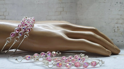 A mannequin hand showcases the Alexa Martha Designs Larger Oval Shaped Swarovski Crystal Bar Bangle Bracelet, alongside silver bangles with pink and iridescent beads, on a light wooden surface and white brick wall background. More Swarovski bracelets are in front.