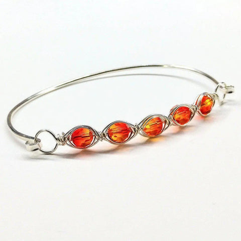 The Larger Oval Shaped Swarovski Crystal Bar Bangle Bracelet by Alexa Martha Designs is a sterling silver bracelet for 8-inch wrists, featuring five orange Swarovski crystals wire wrapped along the band, shown on a white background.