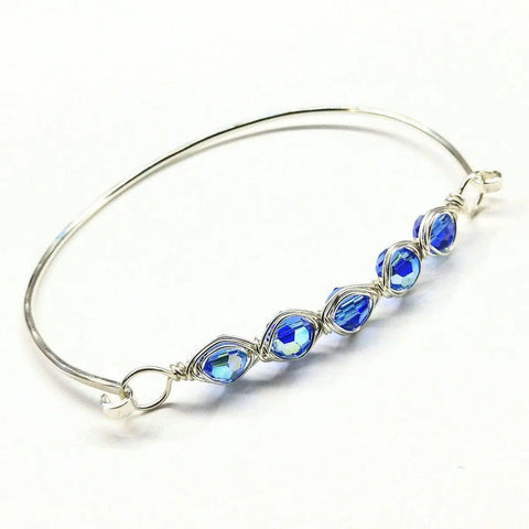 The Larger Oval Shaped Swarovski Crystal Bar Bangle Bracelet by Alexa Martha Designs features sparkling blue faceted beads, wire wrapped for elegance and beautifully displayed on a white background.