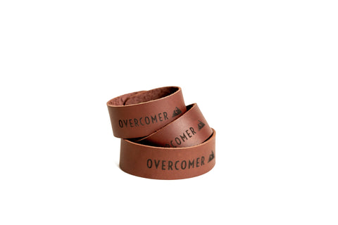The OVERCOMER Cuff Bracelet by Urban Southern is a brown leather bracelet featuring OVERCOMER and a mountain symbol in black, displayed coiled on a white background.