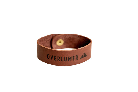 The Urban Southern OVERCOMER Cuff Bracelet is a brown American-made leather cuff with a brass snap and black mountain graphic, shown on a white background.