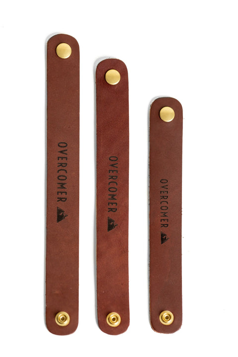 Three Urban Southern OVERCOMER Cuff Bracelets, each brown leather with brass snap buttons and “OVERCOMER” stamped beside a mountain, are arranged vertically on a white background.