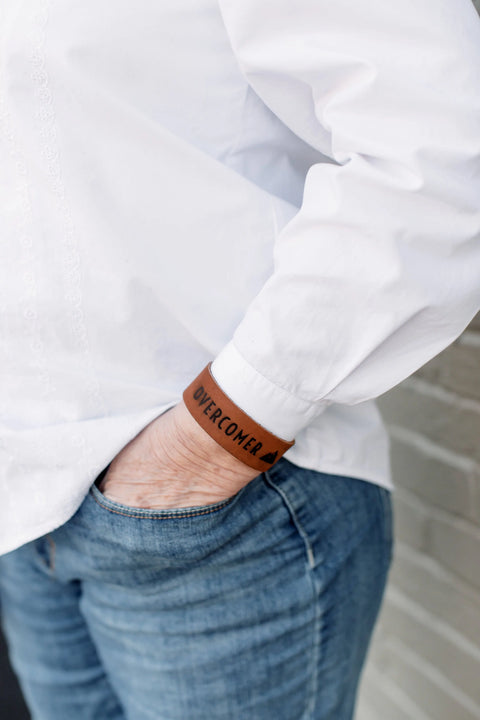 A person in a white shirt and blue jeans has one hand in their pocket, wearing the Urban Southern OVERCOMER Cuff Bracelet—an American-made leather accessory engraved with OVERCOMER for daily inspiration.