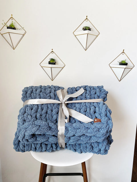 The Comfy Sunday Chunky Knit Blanket in Slate, tied with a white ribbon, rests on a white chair. Behind it, a white wall displays four diamond shelves, each holding a small potted plant.