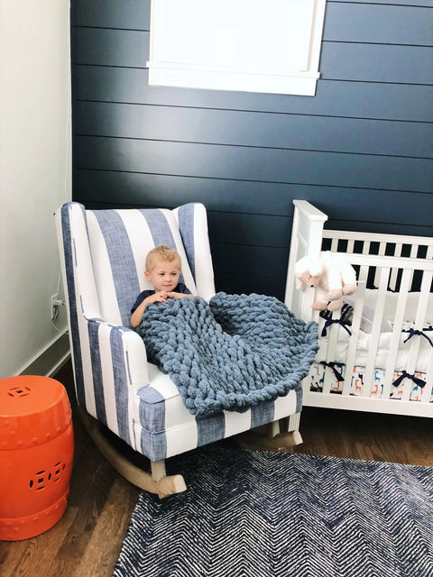 A young child sits in a blue and white striped chair covered with the Comfy Sunday Chunky Knit Blanket in Slate, next to a white crib in a cozy modern nursery with dark blue paneled walls and hardwood floors.