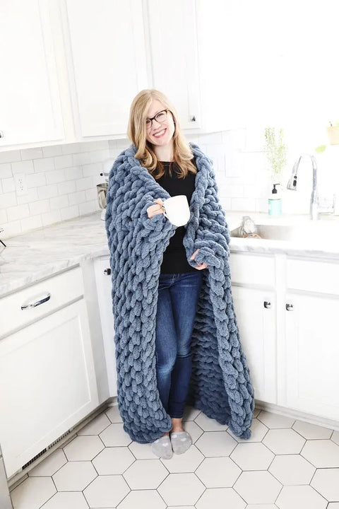 A smiling woman with glasses stands in a white kitchen, wrapped in the Comfy Sunday Chunky Knit Blanket in Slate and holding a white mug. She wears jeans, a black shirt, and cozy gray slippers.