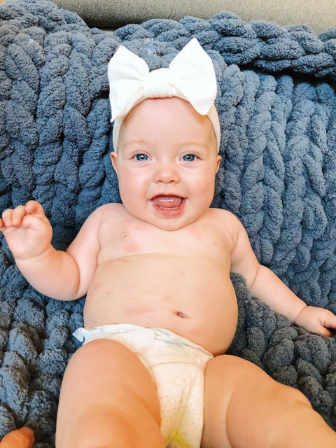 A smiling baby wearing a white bow headband and diaper relaxes on the Comfy Sunday Chunky Knit Blanket in Slate, looking happy and cozy.