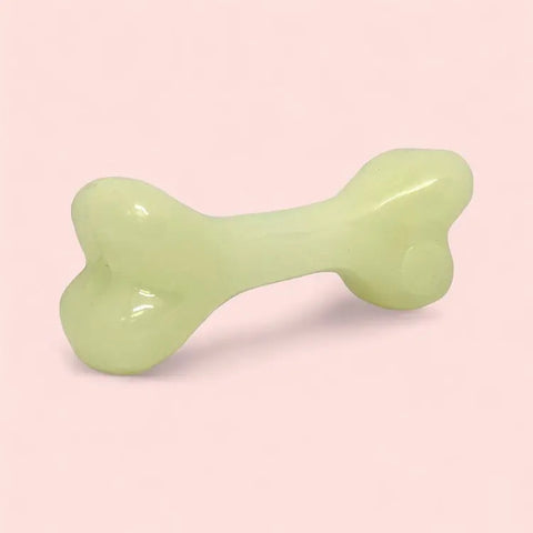 The SodaPup PUP-X Synthetic Rubber Chewing & Floating Bone Toy - Glow in the Dark, a pale yellow-green dog bone-shaped toy, is displayed on a light pink background.