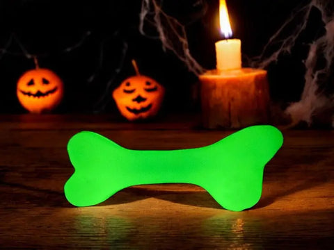 A glowing green SodaPup PUP-X Synthetic Rubber Chewing & Floating Bone Toy - Glow in the Dark rests on a wooden surface, with a candle and two jack-o’-lanterns behind it for a spooky Halloween vibe.