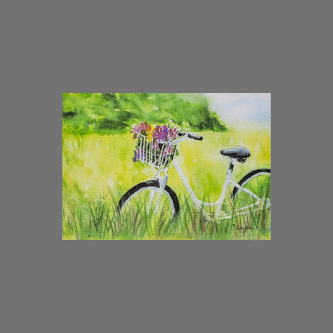 The JuLu Cards Pack of 8 - Bike in the Grass With Flowers (10102) features a white bicycle with a basket of colorful flowers in tall grass, perfect for charming greeting cards set against a vibrant green field and sky.