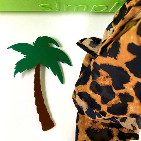 A Chatelet Manufacturing BOGLETS Simply Southern BEACH Palm Tree Charm, in green and brown plastic, is shown next to leopard print fabric and a green bag on a white surface.